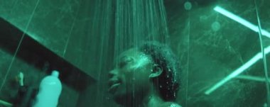 Genç siyahi bir kadının duş kabininde duş alması ve evdeki banyoda yeşil ışıkta durması.