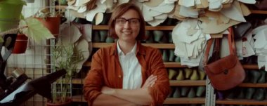 Kafkas kadının gülümseyen ve kameraya poz veren portre fotoğrafını yakınlaştırın. Kolları rafların önünde çapraz dursun. Ayakkabı yapımı atölyesinde kalıplar ve kalıplar devam etsin.