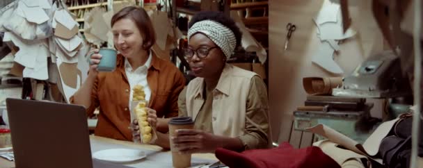 非裔美国人和白人妇女一边吃糕点 一边喝咖啡 一边在笔记本电脑上讨论一些事情 一边在制鞋作坊上谈笑风生 — 图库视频影像