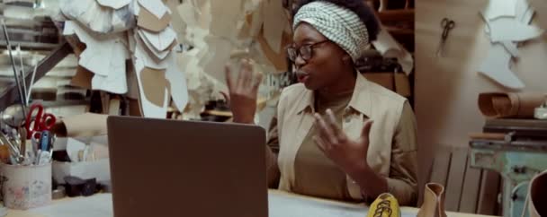 年轻的非洲裔美国妇女坐在制鞋工场的办公桌前 在笔记本电脑上讨论手工制作的鞋子 — 图库视频影像