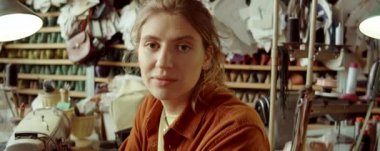 Genç beyaz kadın portresi dikiş makinesiyle masada oturuyor ve atölyede çalışırken kameraya poz veriyor.