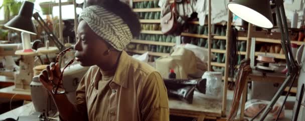 Mujer Afroamericana Reflexiva Sentada Lugar Trabajo Taller Zapateros Sosteniendo Gafas — Vídeos de Stock