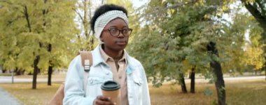 Genç Afrikalı Amerikalı kadın elinde kahve fincanı ile parkta yürüyor.