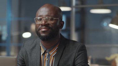 Ofiste geç saatlere kadar çalışırken kameraya bakıp gülümseyen neşeli Afro-Amerikan işadamının portresi.