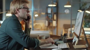 Beyaz erkek mimar telefonda konuşuyor, bilgisayardaki kat planına bakıyor ve ofiste geç saatlere kadar çalışırken notlar alıyor.