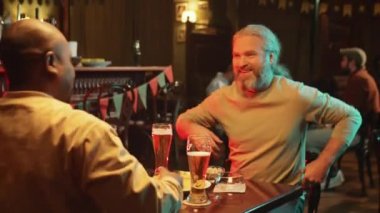 Uzun gri saçlı, olgun beyaz bir adam modern barda Afrikalı Amerikalı arkadaşıyla buluşup onu selamlıyor ve masada oturuyor.