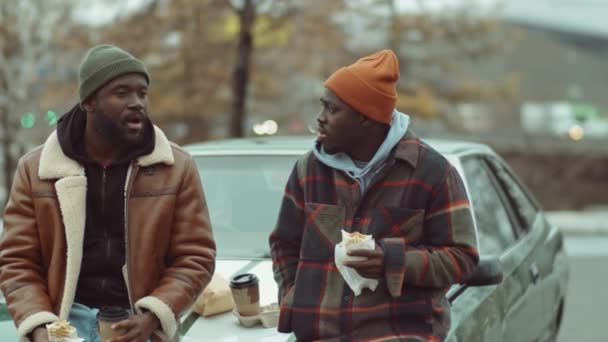 アフリカ系アメリカ人の男性２人が車で外に立ってサンドイッチを食べたりコーヒーを飲んだり — ストック動画