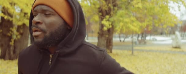 秋の日にアフリカ系アメリカ人の男性が公園でジョギングしているのを胸に撮り 休憩し 周りを見て呼吸する — ストック動画