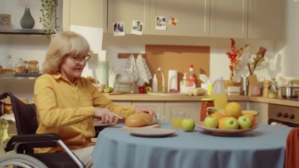 障害のある現代のシニア女性の水平媒体の肖像車椅子に座って朝食のためのキッチンカットパンでテーブル — ストック動画