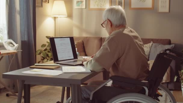 家のノートパソコンの前にある車椅子に座って眼鏡をかけて仕事をする現代のシニアマネージャーの横中出し — ストック動画