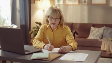 Özürlü, sarı tişörtlü, tekerlekli sandalyede oturan, evinde evrak işleriyle uğraşan, orta yaşlı bir kadının yatay fotoğrafı.