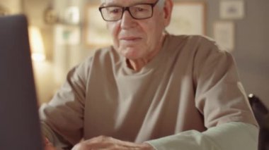 Gözlüklü yaşlı bir adamın yatay orta boy portresi. Oturma odasında masasında oturuyor ve serbest çalışıyor.