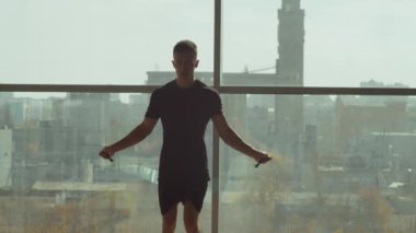 Spor giyinen atletik bir adam spor salonunda panoramik pencerelerle egzersiz yaparken ip atlıyor.