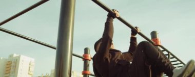 Spor kıyafetli güçlü bir adamın açık hava antrenmanı sırasında yatay barda kol hareketi yaptığı bir yay çekimi.