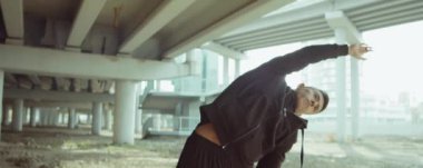 Spor kıyafetleriyle Asyalı sporcunun şehir köprüsünün altında eğildiği ve dışarıda antrenman yapmadan önce ısındığı bir sahne.