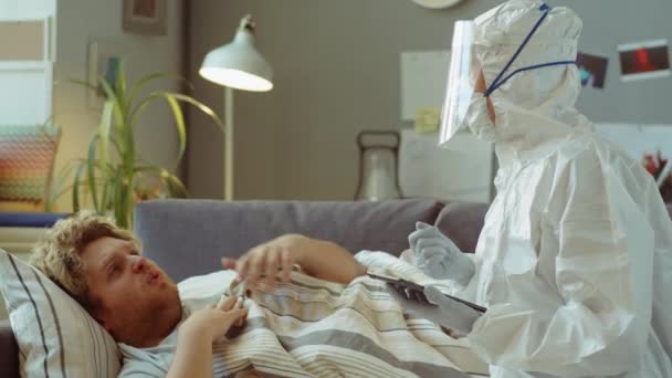 医生身穿防护服 戴着手套 戴着面罩 躺在沙发上 躺在毯子下和使用数码平板电脑与病人交谈 — 图库视频影像