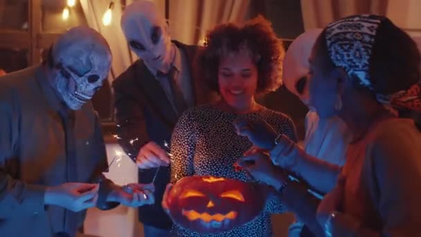 Lächelnde Frau mit Jack-o-Laterne, während ihre Freunde in Halloween-Masken Wunderkerzen mit Kerze anzünden und zu Hause Party tanzen