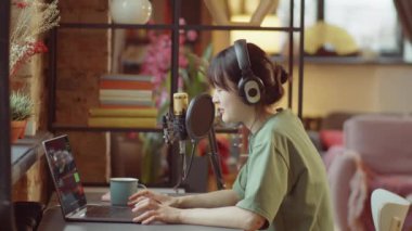 Kulaklıklı genç Asyalı kadın, evde podcast yaparken dizüstü bilgisayar kullanıyor ve mikrofonla konuşuyor.