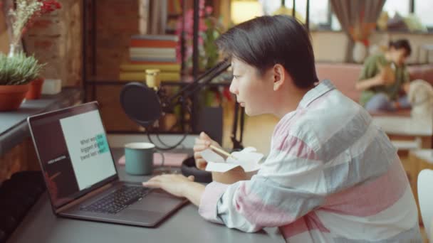 若いアジア人女性が机の上に腰を下ろして 箸で食器棚から鍋を食べ ノートパソコンで作業する — ストック動画
