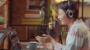 Kulaklık takmış Asyalı bir kadın, evinde podcast yaparken dizüstü bilgisayarla oturuyor ve mikrofonla konuşuyor.