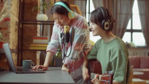 两名戴耳机的年轻亚洲妇女在家里一起录音时使用笔记本电脑和吉他 — 图库视频影像