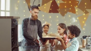 Neşeli çoklu etnik çocuklar gülümsüyor ve aşçıya yemek çarşaflarını fırına koyması için yardım ediyor.