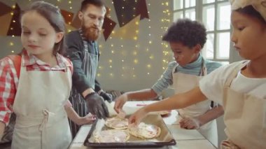 Yemek kursunda erkek şefin yardımıyla pişirmeden önce pizzaya jambon ekleyen bir grup çocuk.