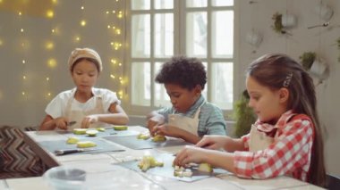 Küçük çoklu etnik kızlar ve oğlan mutfak masasında taze armut kesiyorlar ve ana yemek kursu sırasında konuşuyorlar.