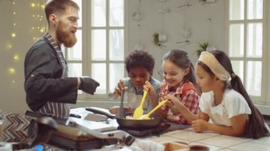 Aşçılık dersi sırasında aşçıbaşıyla yemek pişirirken tavada yemek karıştıran çok ırklı küçük çocuklar.