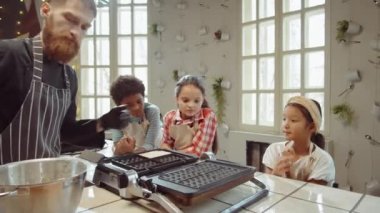 Küçük kız waffle makinesine hamur dökerken aşçı ve diğer çocuklar yemek yaparken onu izliyor.
