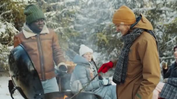 年轻的白人男子在烧烤烧烤中转炭 并与非洲裔美国朋友聊天 因为他吃烤棉花糖在森林冬季野餐 — 图库视频影像