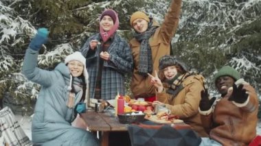 Kış ormanlarında piknikte kamp masasında otururken kameraya gülümseyen ve el sallayan çok ırklı genç arkadaşların toplu portresi.