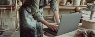 Çömlek atölyesinde dizüstü bilgisayar kullanan beyaz önlüklü genç bir kadını yukarı kaldır.