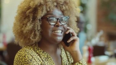 Gözlüklü genç Afrikalı Amerikalı kadın kafede otururken cep telefonuyla bir şey tartışıyor.