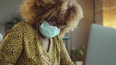 Koruyucu maskeli ve gözlüklü Afrikalı Amerikalı iş kadını not alıyor ve covid-19 pandemik zamanlarda ofiste çalışırken masamda dizüstü bilgisayar kullanıyor.