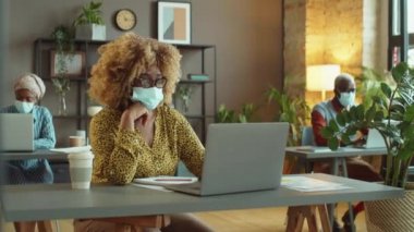 Koruyucu maskeli Afro-Amerikalı iş kadını covid-19 pandemik zamanlarda ofiste çalışırken dizüstü bilgisayarında yazı yazıyor.