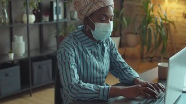 Korunaklı maskeli Afrikalı Amerikalı iş kadınının Coronavirus salgını sırasında ofis masasında dizüstü bilgisayarla çalışırken görüntüsünü kaldır.