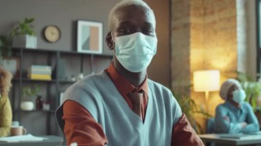Korumacı yüz maskeli Afrikalı Amerikalı iş adamının portresi ofiste oturmuş Coronavirus salgını sırasında kameraya poz veriyor.