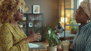 Afrika kökenli Amerikalı iş kadını kahve fincanı ile ayakta duruyor ve ofis işi sırasında bayan meslektaşıyla konuşuyor.