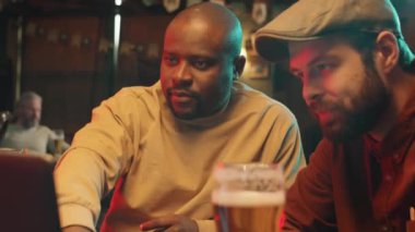 Orta boy Afrika kökenli Amerikalı ve Kafkasyalı erkeklerin birlikte oturup bira içerken ve dizüstü bilgisayar kullanırken çekilmiş fotoğrafları.