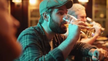 Yüzünde sakalı olan genç beyaz bir adam modern barda arkadaşlarıyla masaya oturmuş nefis bira ve sohbetin tadını çıkarıyor.