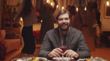 Yemek masasında oturan yakışıklı genç adamın yatay yavaş portresi. Kameraya gülümsüyor, arkadaşları arkasında dans ediyor.