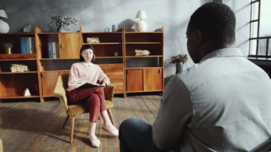Genç, güzel bir kadın psikoloğun omuz üstü fotoğrafı elinde dosyayla koltukta oturuyor ve danışmanlık sırasında ona danışmanlık yaparken Afrikalı Amerikalı erkek hastayla konuşuyor.