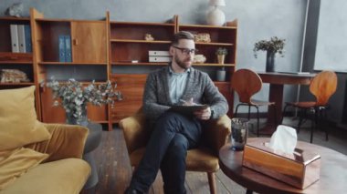 Ofisindeki koltukta oturan beyaz erkek psikoloğun görüntüsünü yakınlaştır. Not alıyor ve danışmanlık ofisinde çalışırken kameraya poz veriyor.