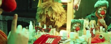 Makyajlı ve kırmızı burunlu palyaçonun aynadaki yansıması soyunma odasında oturuyor ve surat yapıyor.