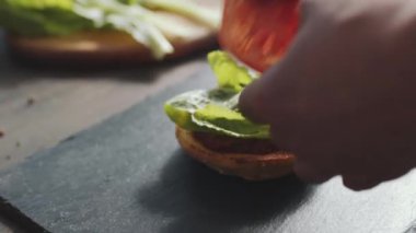 Hamburger hazırlarken, erkek ellerinin çöreğe salatayla domates dilimleri kattığı yakın çekim.