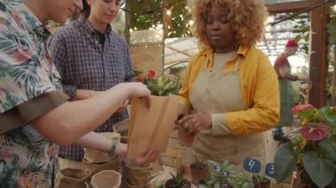 Afro-Amerikalı satıcı kağıt torbaya saksı koyuyor ve çiçek pazarında neşeli beyaz kadın müşterilerle konuşuyor.