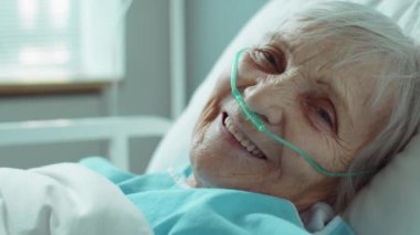 Hastane koğuşunda yatağında yatıp kameraya gülümseyen burun kanülü pozitif yaşlı kadın portresi.