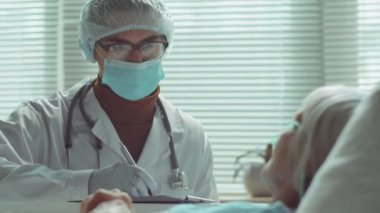 Yaşlı kadın hastanın hastane koğuşunda yatarken ve koruyucu maske takmış doktorla konuşurken görüntüsünü kaldır.