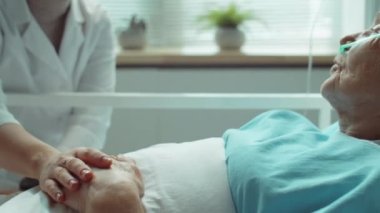Genç bayan doktor, yaşlı kadının elini tutuyor, hastanede terapi sırasında gülümsüyor ve onunla konuşuyor.